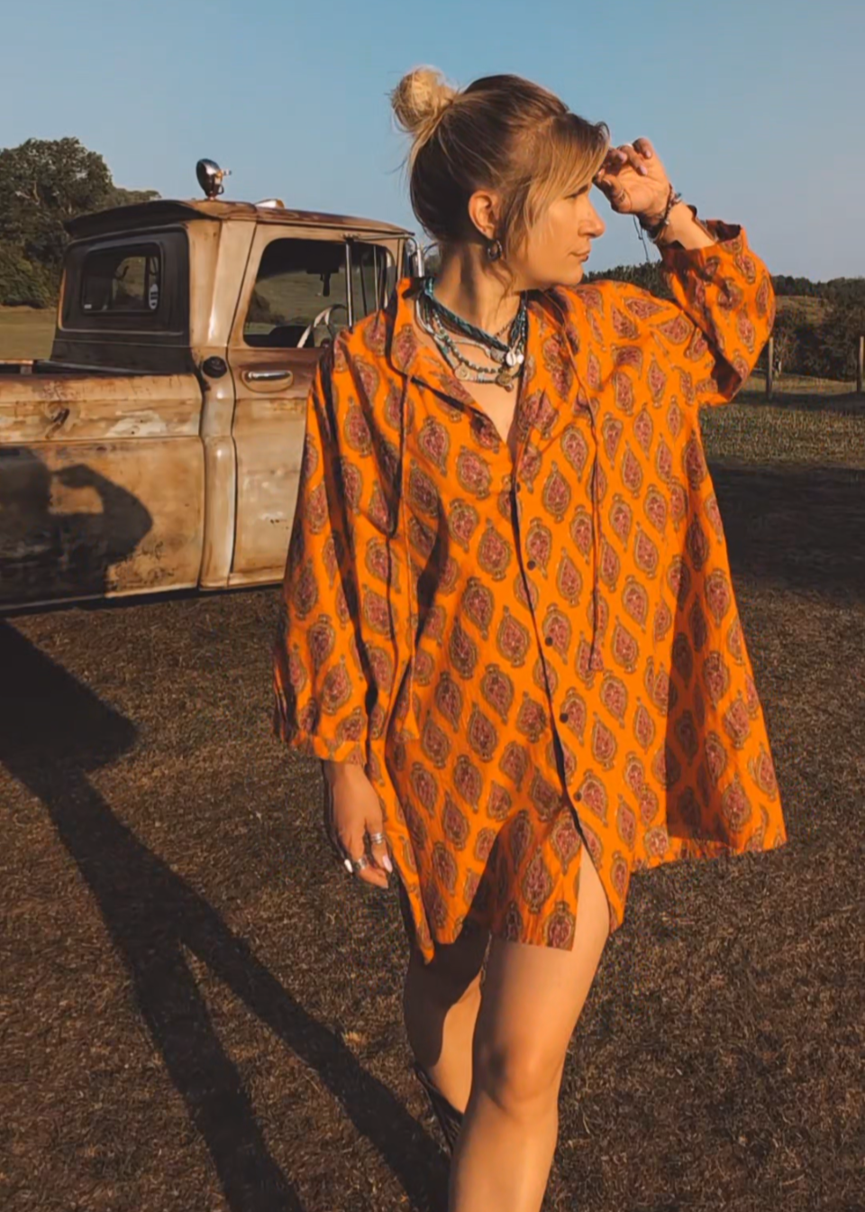 Woman in an orange patterned dress standing in a field with a vintage truck in the background.
